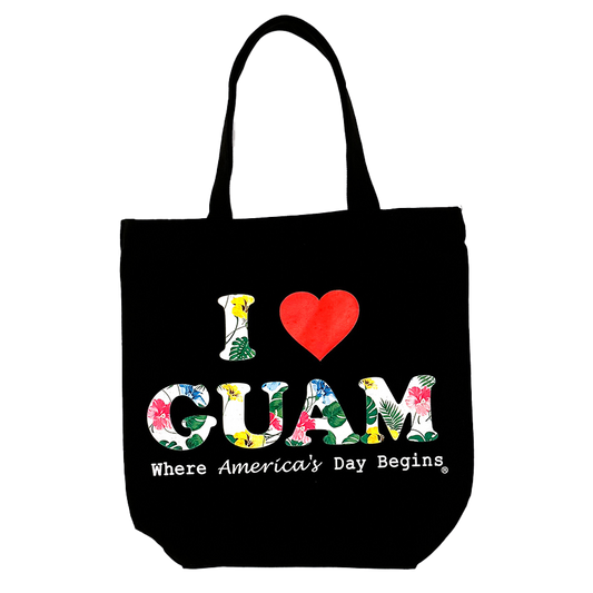 Black canvas tote bag featuring the ‘I ❤️ GUAM’ design with floral-patterned letters and the slogan ‘Where America’s Day Begins’ printed in white beneath the logo.