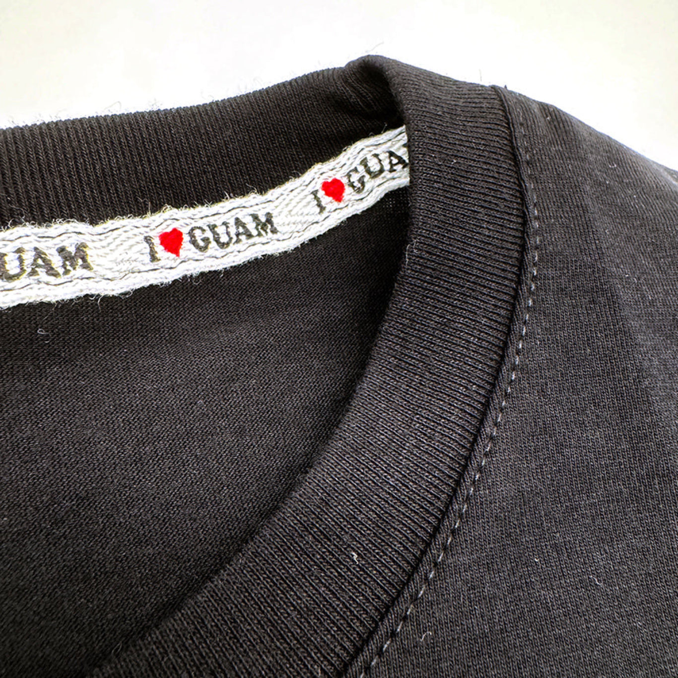 Close-up view of the neckline of a black ‘I ❤️ GUAM’ unisex t-shirt, featuring woven interior branding with repeated ‘I ❤️ GUAM’ text along the inside collar.