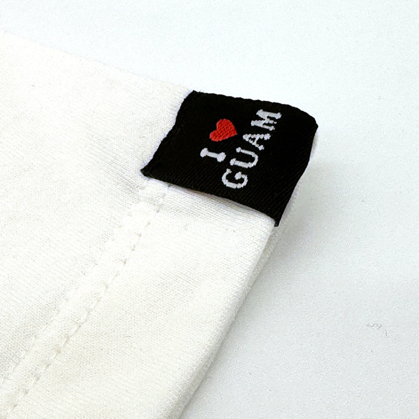 Close-up of the sleeve hem of a white ‘I ❤️ GUAM’ unisex t-shirt, featuring a black woven label with the ‘I ❤️ GUAM’ logo stitched in white and red.